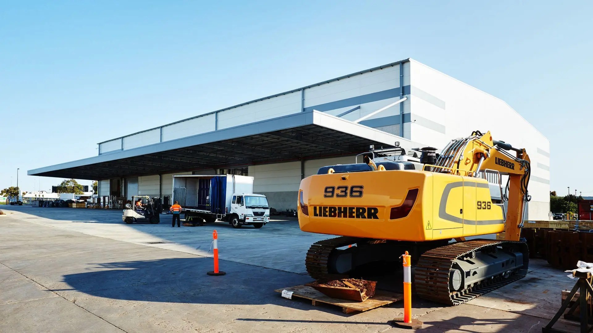 Excavator parked on a concrete yard at an industrial depot, with a delivery truck and loading dock